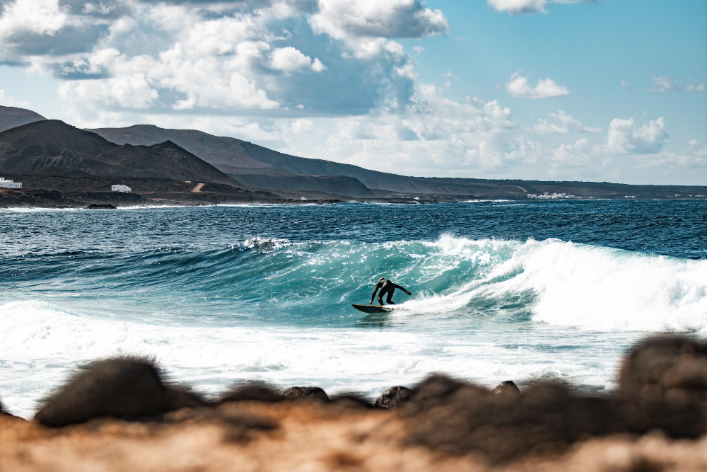 Surfer auf einer Welle vor der felsigen Küste von Fuerteventura – ideales Ziel zum Wellenreiten auf den Kanaren