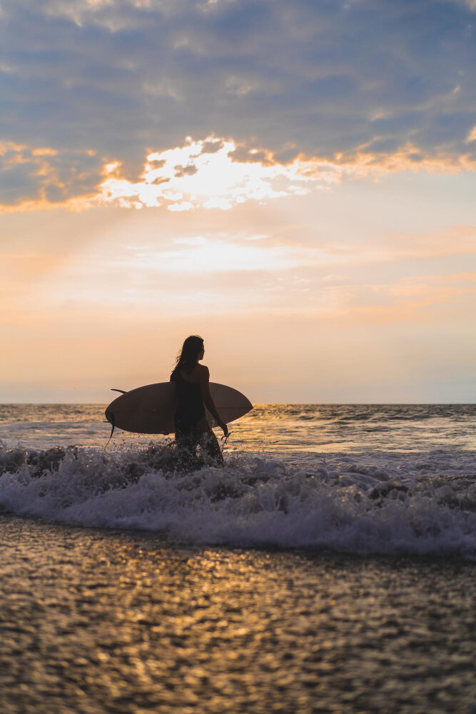 Surferin mit Surfboard läuft bei Sonnenuntergang ins Meer – Surfen in Marokko am Atlantik