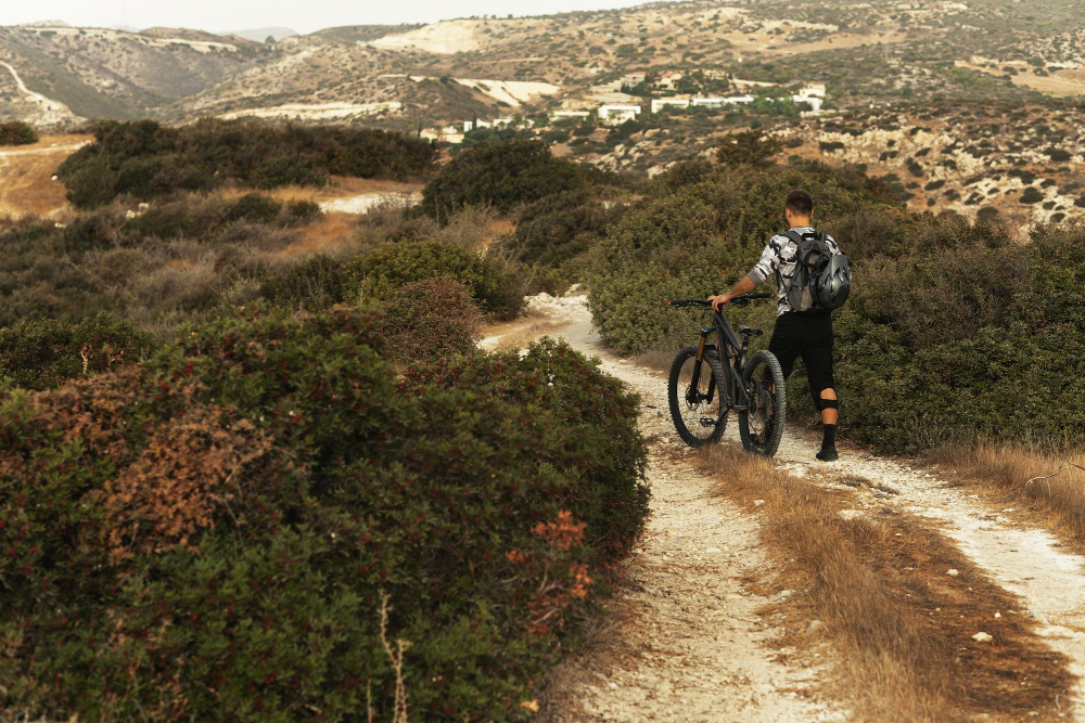 Mountainbiker schiebt sein Rad auf einem mediterranen Trail in Portugal, umgeben von Sträuchern und Hügeln – Outdoor-Abenteuer abseits der Küste