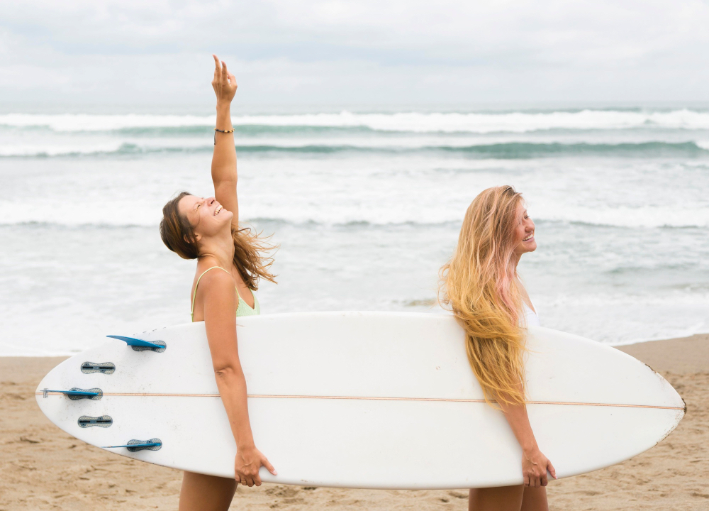 Zwei fröhliche Surferinnen tragen gemeinsam ein Surfboard am Strand und genießen die Wellen in Portugal.