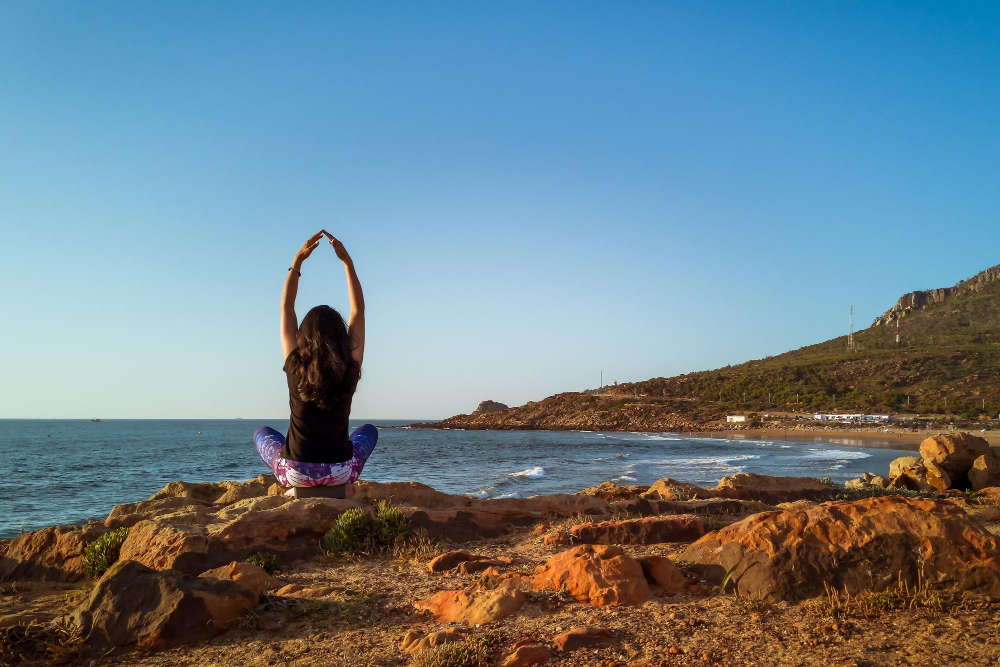 Frau praktiziert Yoga am Meer in Portugal, sitzend auf Felsen mit Blick auf den Ozean – Entspannung nach dem Surfen