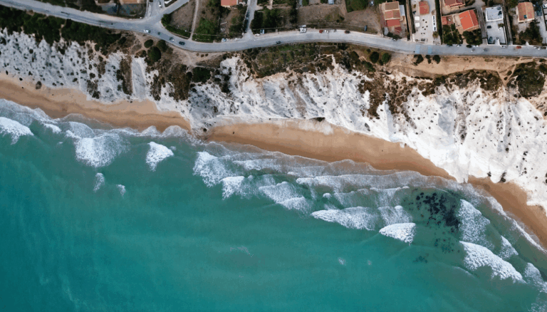 Luftaufnahme einer goldenen Sandküste in Spanien mit türkisblauem Meer und sanften Wellen, eingerahmt von hellen Klippen und Küstenhäusern.
