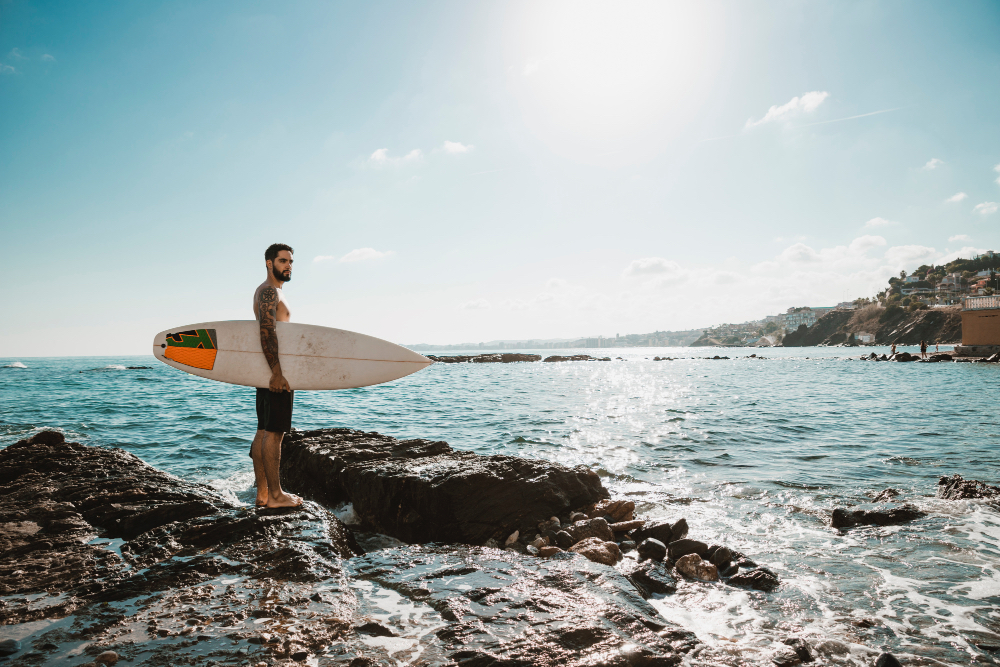Surfer mit Board auf den Lavaklippen an der Südküste Teneriffas mit Blick auf das Meer und die Sonne über der Atlantikküste.