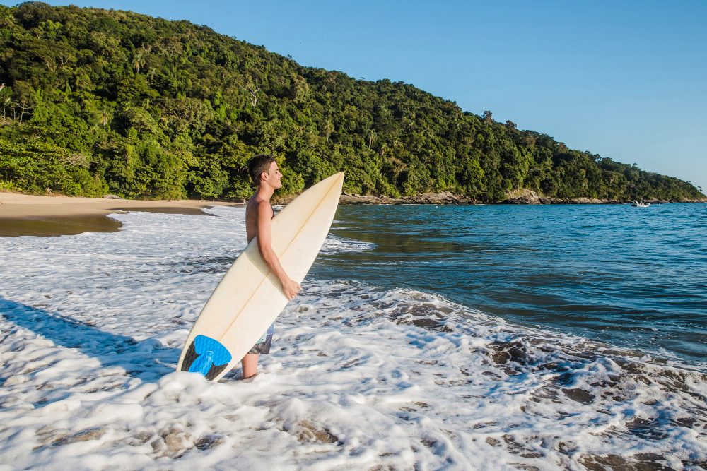 Surfer mit Board steht im Schaum der Wellen an einem tropischen Strand in Sri Lanka mit dichtem Dschungel im Hintergrund